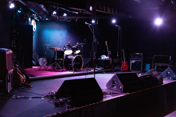 Leicht körniges Foto der Bühne im Blues Club Meisenfrei. Das licht hat einen mystischen violetten und hellblauen Touch. Auf der Bühne stehen Schlagzeug, Bass und Gitarren, sowie Mikrophone der Band bereit um heute Nacht als Mad Dogs Blues Band Bremen so richtig krass zu rocken.