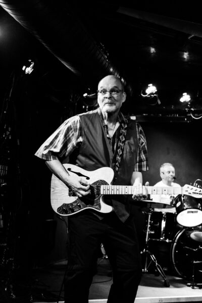 Ein lustiges Foto eines Gitarristen im Blues Club Meisenfrei. Es ist schwarz weiß und er guckt hoch konzentriert an der Kamera vorbei. Wobei er sich nicht entscheiden kann ob er ein konzentriertes oder lustiges Gesicht machen soll und sich auf dem Weg dort hin irgendwie verrant hat. Leider kam in dem Moment die beste Fotografin Bremens, die Photonenhexe und hat genau in dem Moment auf den Auslöser geklickt.