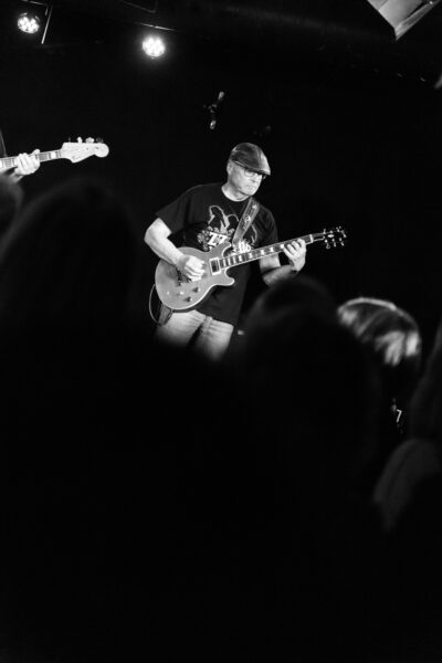 Ein legendäres Schwarz Weiß Foto vom Gitarristen der Band Mad Dogs Blues Band. Im Vorderrgrund verwischt das Publikum zu einer schwarzen Wand. Während der Musiker im Blues Club Meisenfrei klar zu sehen ist wie er einen besonders schweren Akkord spielt.