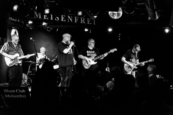 Die gesammte Band Mad Dogs Blues Band im Meisenfrei aus der frontalen Fotografiert. Vorne steht Publikum, hinten ist die Band. Der Fotostil ist im harten schwarz weiß Kontrast. Das Foto wirkt Zeitlos.