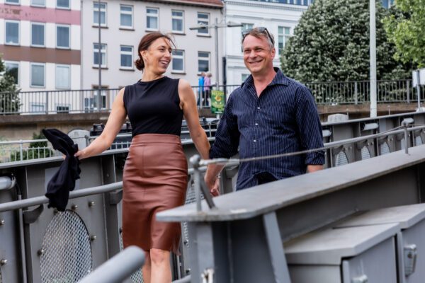 Ein Paar geht lächelnd über eine Metallbrücke in Bremen. Die Frau trägt einen braunen Lederrock und ein schwarzes Top, der Mann ein dunkelblaues Hemd. Beide halten sich an den Händen und schauen sich mit offenem Lachen an. Die Szene wirkt lebendig, ungestellt und voller gemeinsamer Freude. Im Hintergrund sind Bäume, Gebäude und Passant:innen zu sehen.