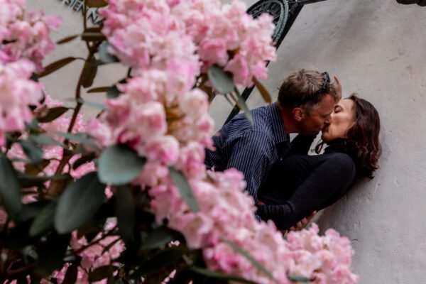Ein Paar küsst sich innig an einer hellen Wand, halb verborgen hinter rosa blühenden Rhododendronzweigen. Die Frau lehnt rücklings an der Wand, während der Mann sie zärtlich hält. Beide tragen dunkle Kleidung, das Licht ist weich und natürlich. Der Fokus liegt auf dem Kuss, der von den Blüten im Vordergrund eingerahmt wird.