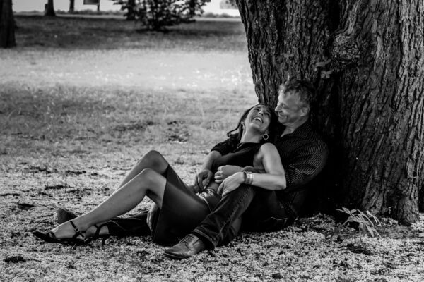 Ein Paar sitzt gemeinsam am Fuß eines großen Baumes im Park. Die Frau lehnt sich lachend zurück in die Arme des Mannes, der hinter ihr sitzt und ebenfalls lächelt. Beide tragen elegante Kleidung, wirken aber vollkommen entspannt. Die Szene ist in Schwarz-Weiß aufgenommen und vermittelt Leichtigkeit, Vertrautheit und echte Freude.